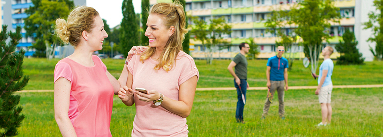 Zwei Frauen stehen im Vordergrund auf einer Wiese und vor einem Wohnblock und Lachen. Im Hintergrund spielen drei Personen Federball.